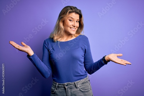Young beautiful blonde woman wearing casual t-shirt over isolated purple background smiling showing both hands open palms, presenting and advertising comparison and balance