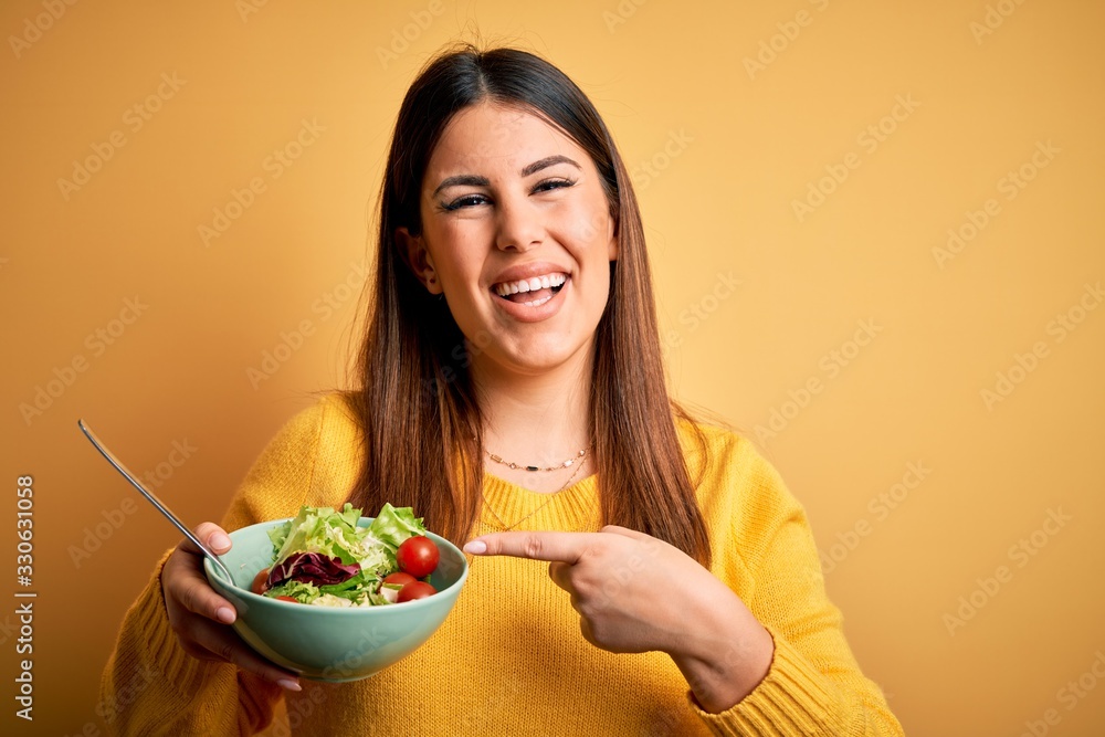 Young beautiful woman eating healthy fresh salad over yellow background very happy pointing with hand and finger