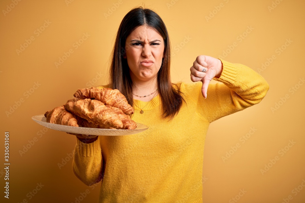Young beautiful woman holding french croissant pastry over yellow background with angry face, negative sign showing dislike with thumbs down, rejection concept Young beautiful woman holding french croissant pastry over yellow background with angry face, negative sign showing dislike with thumbs down, rejection concept