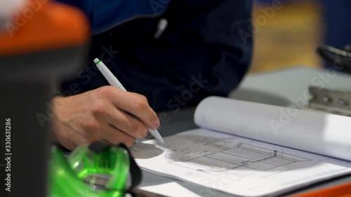 Hand of male builder writes, draws, makes notes in construction project or log book on table next to documents and papers with drawn plan. Close up view. Unrecognizable foreman working.