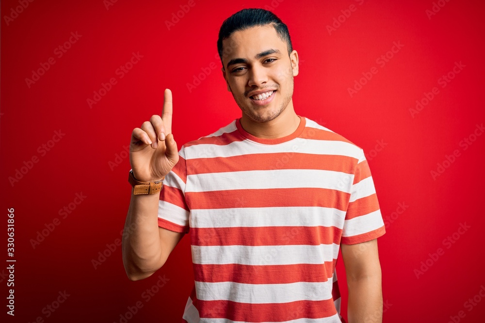 Young brazilian man wearing casual striped t-shirt standing over isolated red background showing and pointing up with finger number one while smiling confident and happy. Young brazilian man wearing casual striped t-shirt standing over isolated red background showing and pointing up with finger number one while smiling confident and happy.