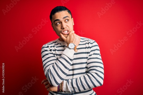 Young brazilian man wearing casual striped t-shirt standing over isolated red background Thinking worried about a question, concerned and nervous with hand on chin