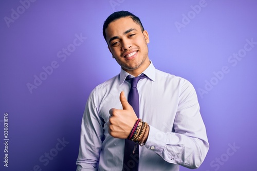 Young brazilian businessman wearing elegant tie standing over isolated purple background doing happy thumbs up gesture with hand. Approving expression looking at the camera showing success.