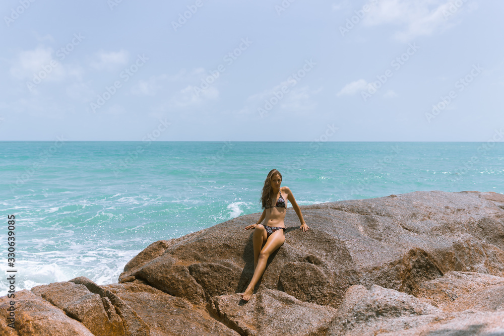 A beautiful girl in a swimsuit is standing on the beach on the rocks with the sea.Against her background,waves and splashes splash.The concept of a traveler and a good life in Thailand.Model