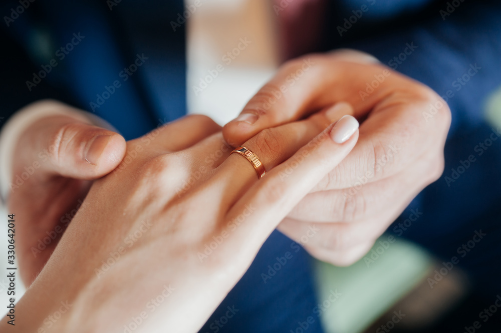 The groom in a blue suit puts a gold ring on the hand of the bride in a white stylish dress. Hands of the newlywed close-up, no faces. The groom in a blue suit puts a gold ring on the hand of the bride in a white stylish dress. Hands of the newlywed close-up, no faces.