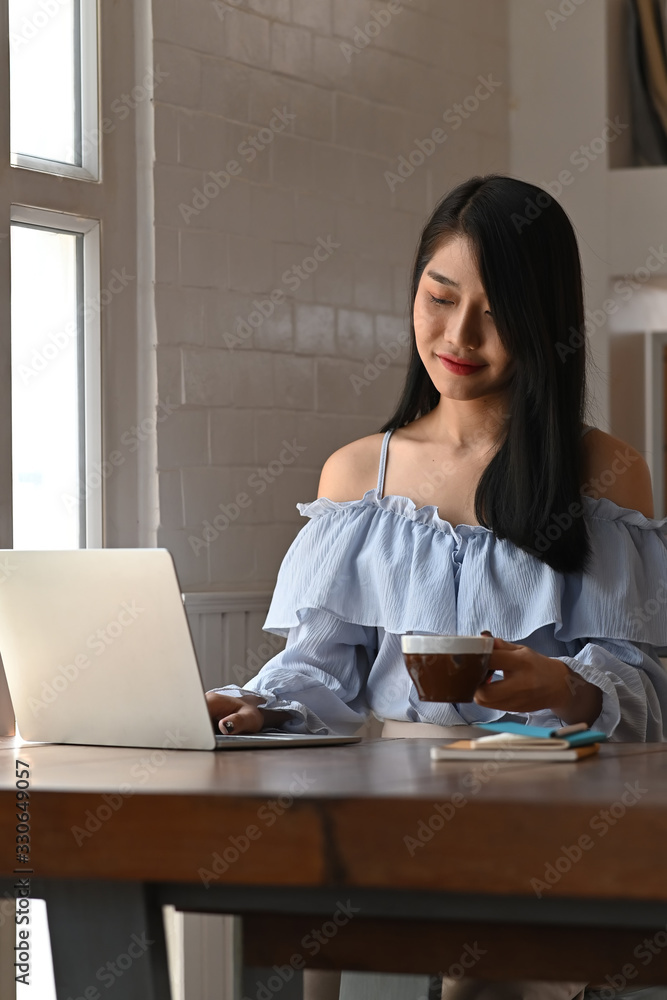 Photo of beautiful woman holding a coffee cup in hand while typing on computer laptop and sitting at the wooden working table over comfortable living room as background. Photo of beautiful woman holding a coffee cup in hand while typing on computer laptop and sitting at the wooden working table over comfortable living room as background.