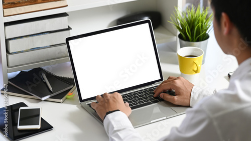 Behind of Young smart man working as Data analyst typing on computer laptop with white blank screen while sitting at the modern working table that surrounded by office equipment.