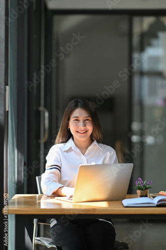 Photo of young beautiful woman smiling while sitting in front her computer laptop at the working wooden table over modern office as background. © Prathankarnpap