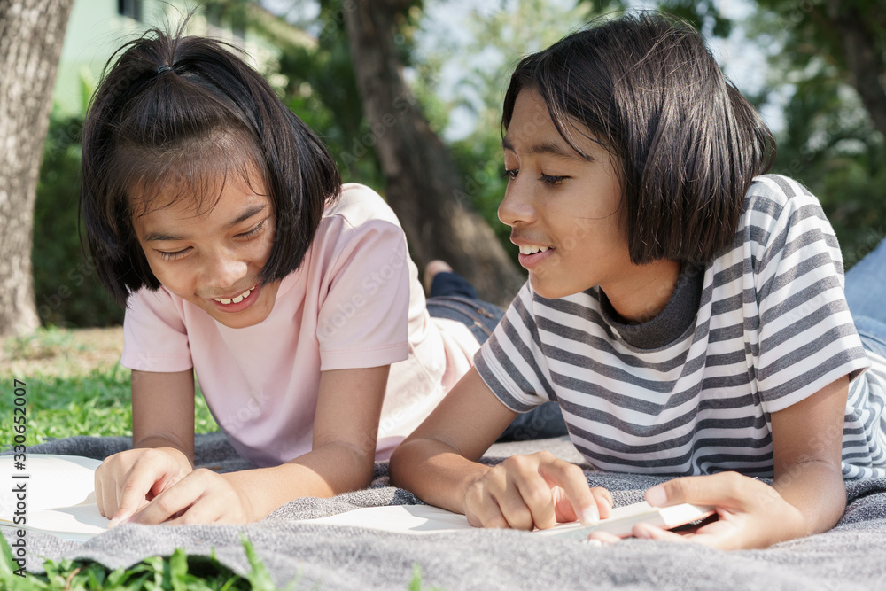 Happy Asian elementary while lying read book on blanket in the summer, Asia two girl smile and relaxation together at park in the morning. The schoolgirl came to study field trip and learned outdoor Happy Asian elementary while lying read book on blanket in the summer, Asia two girl smile and relaxation together at park in the morning. The schoolgirl came to study field trip and learned outdoor