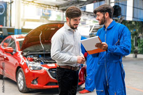 Mechanic show the car checking list for customer with blur his assistant checking red car. Focus on mechanic and customer on the right side. Auto car repair service center. Professional service. photo