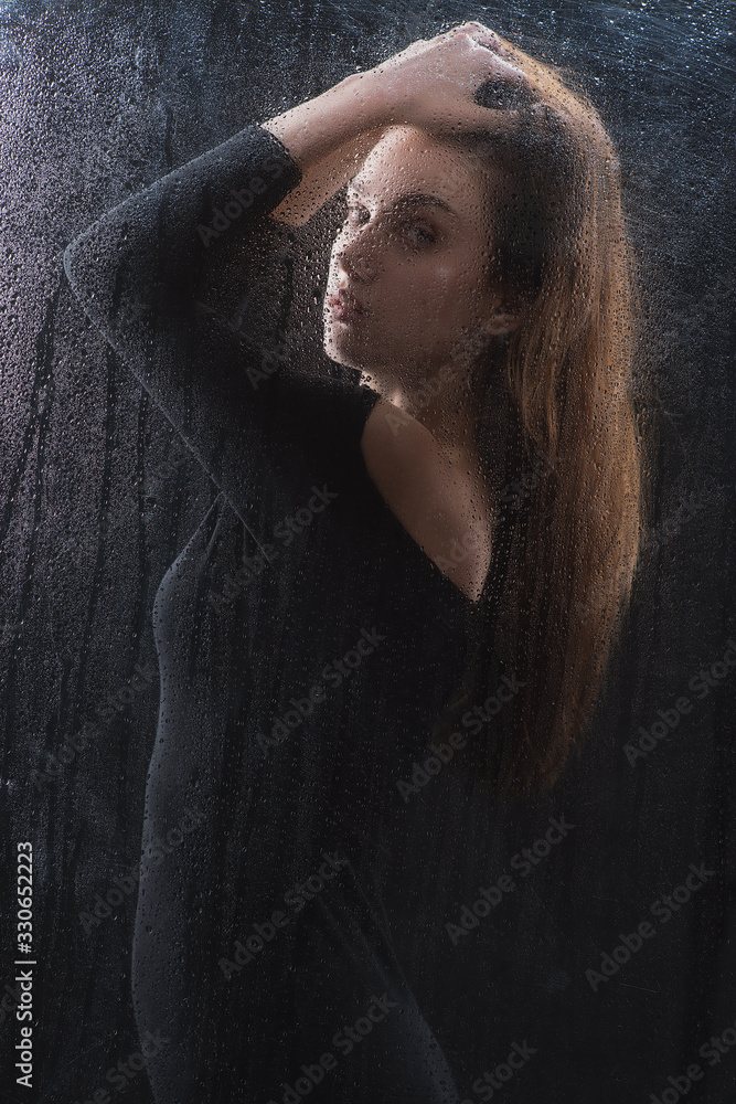Beautiful slender brunette in a black dress with a good figure behind the glass with raindrops, the silhouette of a woman behind a blind glass in the fog