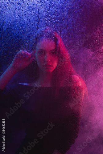 A beautiful slender girl with long hair behind a blind glass in neon lights in a black dress