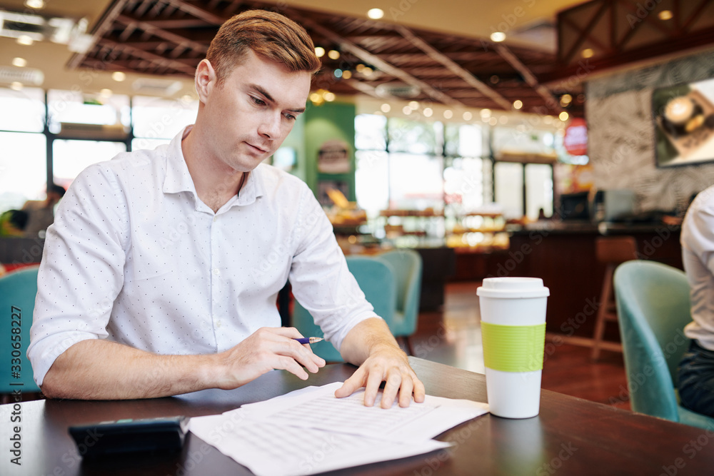 Fototapeta premium Handsome young entrepreneur sitting at cafe table with cup of take away coffee and checking figures in financial report