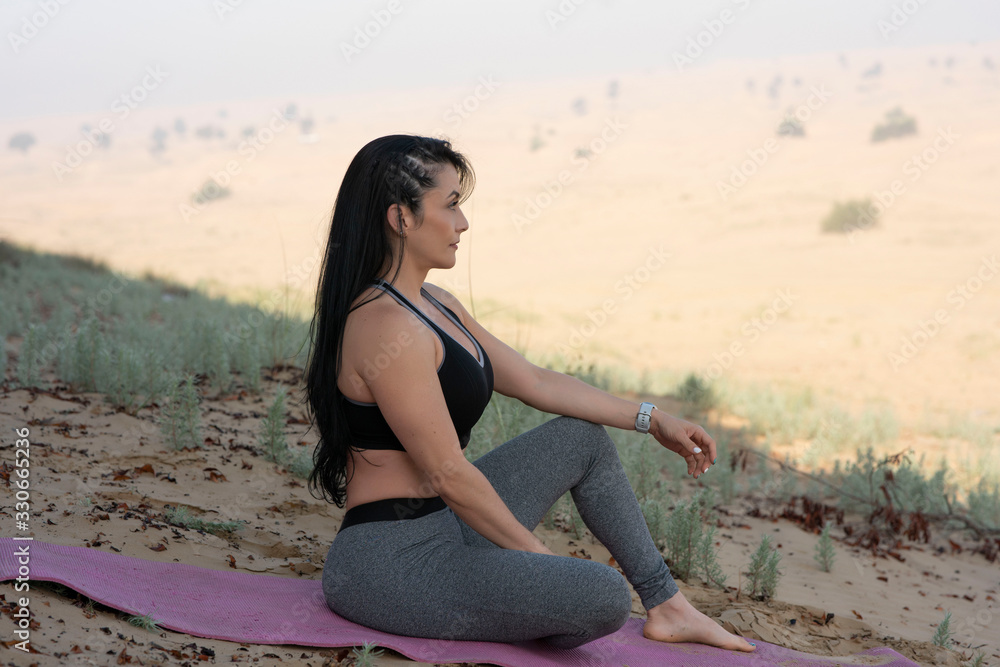 Beautiful long haired brunette hispanic athletic Yogi woman wearing a black sports bra and gray tights sitting on the edge of a desert meditating relaxing thinking on. summer day in the Middle East