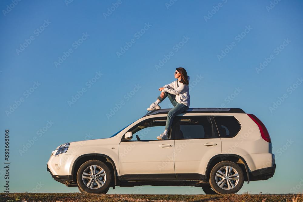 stylish woman in casual outfit sitting on the roof of the car at sunset