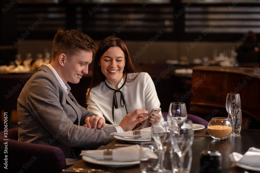 attractive caucasian lady show something interesting on smartphone to boyfriend. guy look at screen of mobile phone and smile, laugh. happy couple in restaurant