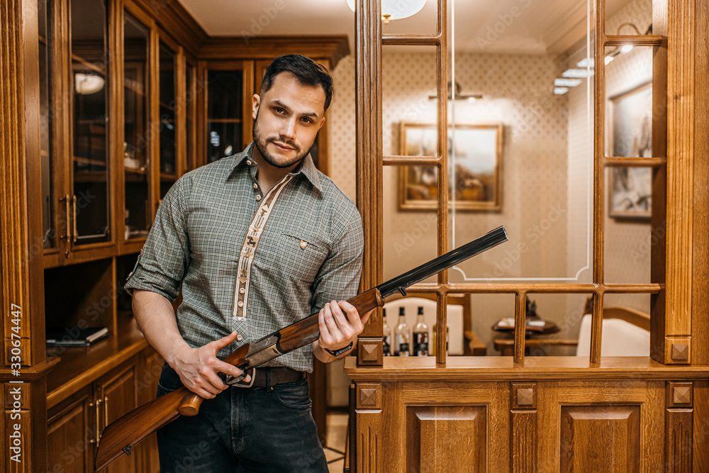 portrait of young caucasian hunter man going to buy new weapon in guns store, he hold rifle in hands, try to use it. indoors, around showcases with rifles portrait of young caucasian hunter man going to buy new weapon in guns store, he hold rifle in hands, try to use it. indoors, around showcases with rifles
