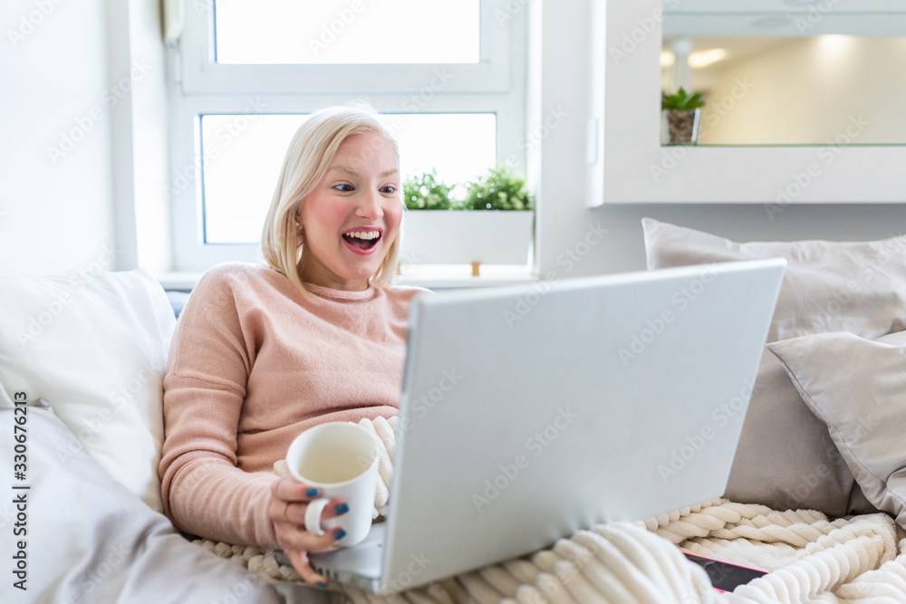 Beautiful young woman drinking coffee at home in her bed wearing a cozy t shirt while working on her laptop. Checking email in the morning, freelance concept Beautiful young woman drinking coffee at home in her bed wearing a cozy t shirt while working on her laptop. Checking email in the morning, freelance concept
