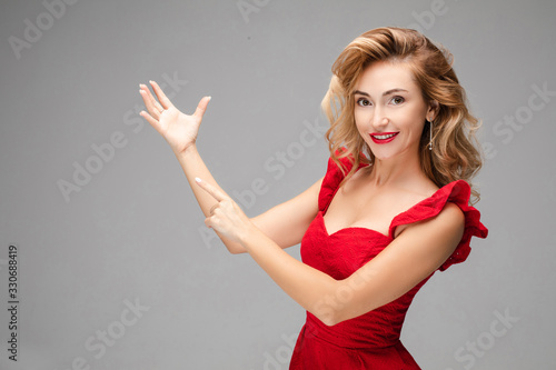 Portrait of cheerful young caucasian female with long blonde hair, bright makeup, red lips in red dress points to her hand