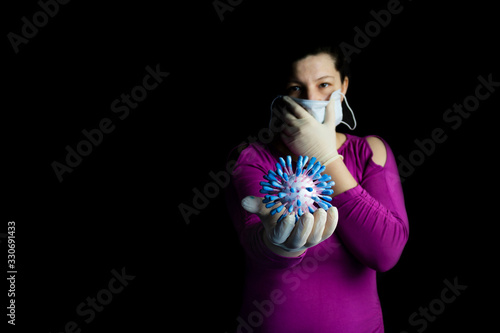 A dark room. In it, a girl with dark hair with one hand holds a medical mask at her mouth. On the other hand is a model of the coronavir molecule photo