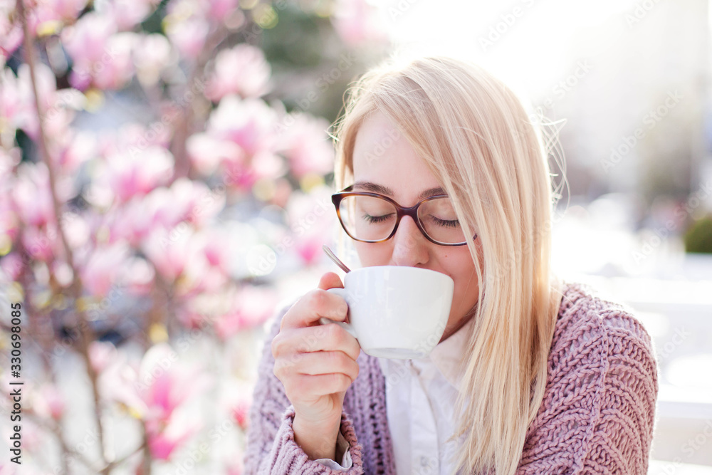 Young woman drinking coffee in spring cafe at city streets. Happy girl enjoying morning breakfast in blooming pink magnolia flowers outdoors. Lifestyle moment. Concept of spring mood, tenderness. Young woman drinking coffee in spring cafe at city streets. Happy girl enjoying morning breakfast in blooming pink magnolia flowers outdoors. Lifestyle moment. Concept of spring mood, tenderness.