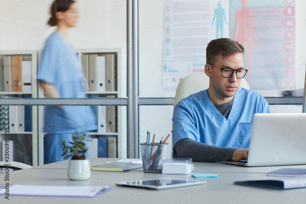 Portrait of male doctor using computer and wearing glasses while sitting at workplace in medical office interior, copy space Portrait of male doctor using computer and wearing glasses while sitting at workplace in medical office interior, copy space