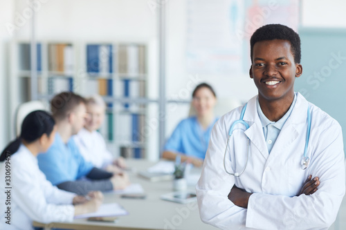 Waist up portrait of young African-American doctor smiling at camera while standing with arms crossed against medical conference background, copy space