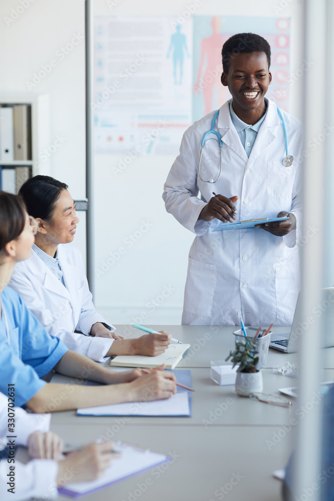 Fototapeta premium Portrait of young African-American doctor giving speech while standing at table during medical council or conference in clinic, copy space