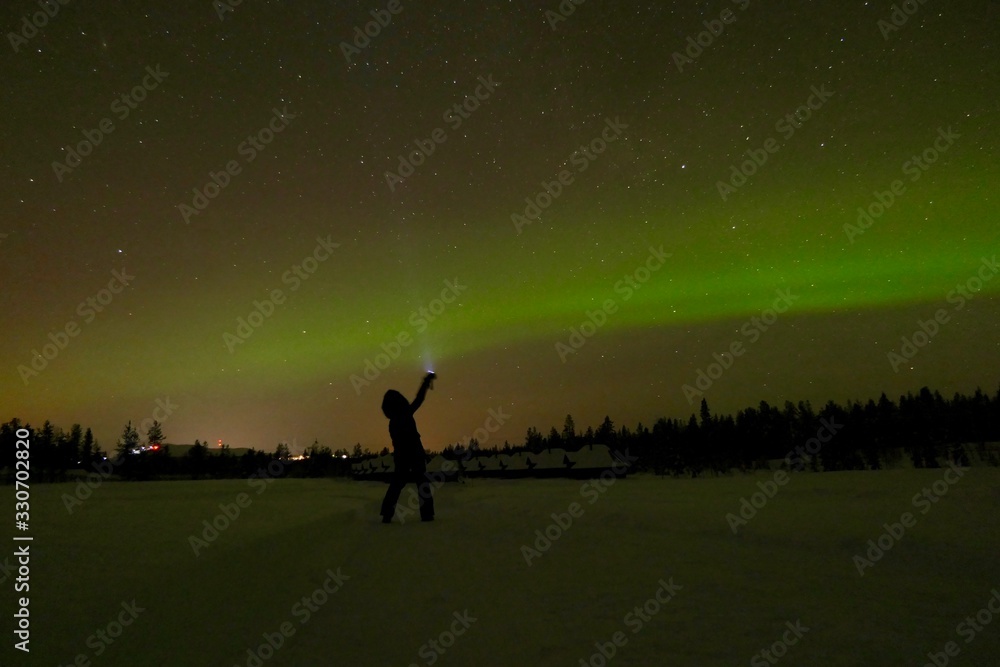 Woman with flashlight before green aurora borealis at night in snow landscape, green and orange sky, cottages in background, Lapland, Finland