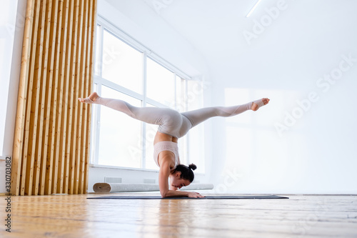 Side view of young caucasian girl gymnast doing pincha mayurasana exercise handstand pose on mat on floor of panoramic window. Concept of professional athletes gymnasts and acrobats. Advertising space photo