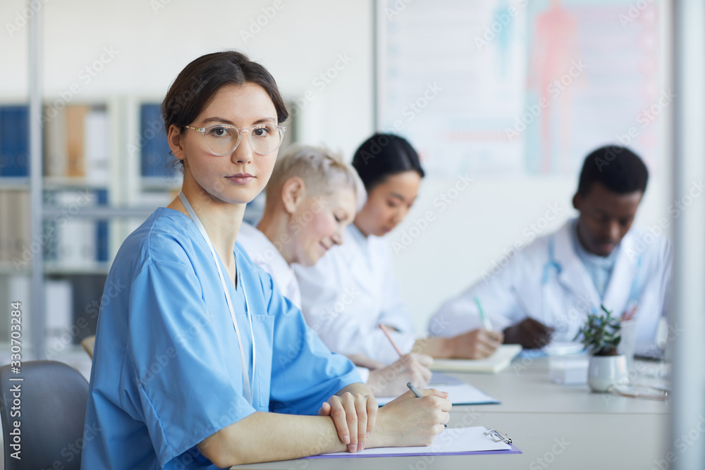 Portrait of young female doctor looking at camera while sitting at table during medical council or conference in clinic, copy space