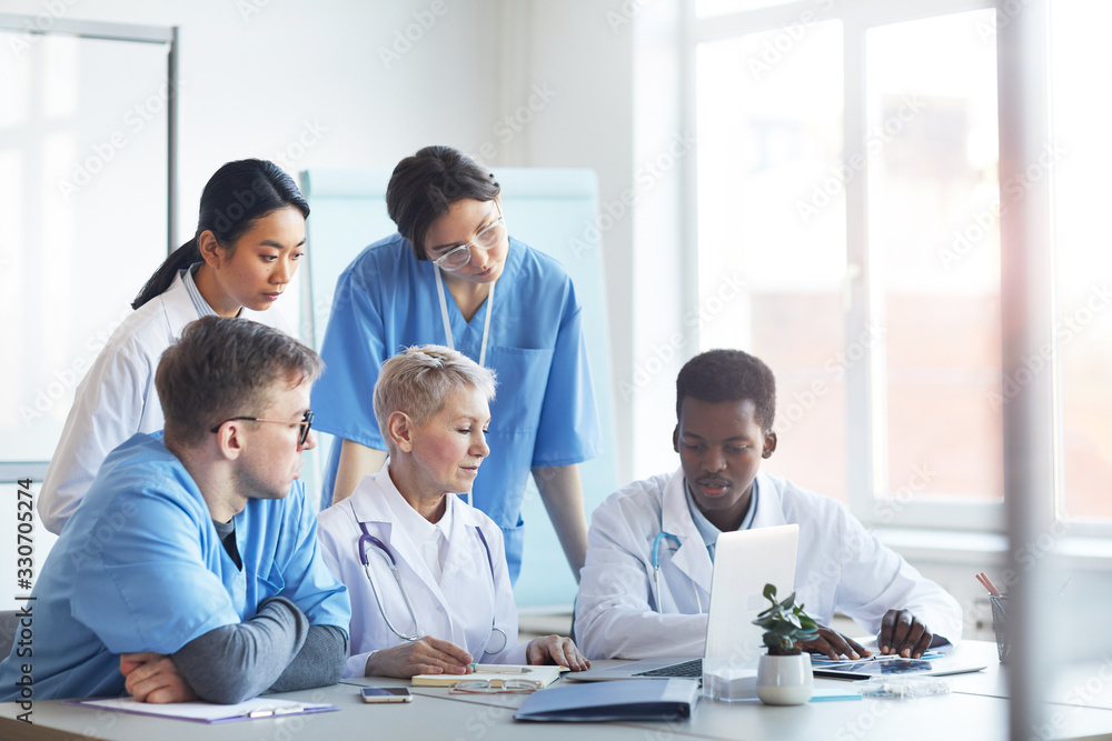 Multi-ethnic group of doctors looking at laptop screen while working together sitting at desk in medical office interior Multi-ethnic group of doctors looking at laptop screen while working together sitting at desk in medical office interior
