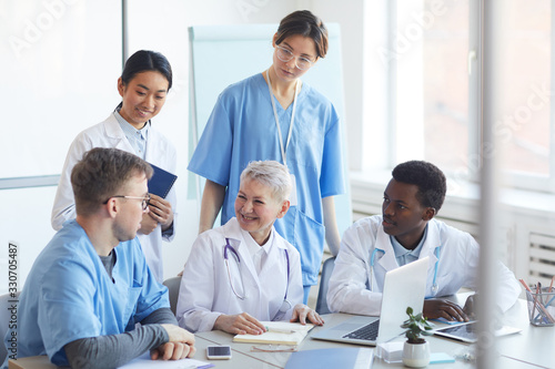 Multi-ethnic group of doctors smiling cheerfully while working together sitting at desk in medical office interior