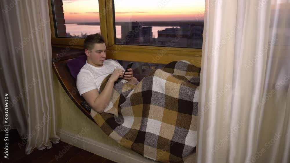 A young man is texting on the phone. A man lies on a window sill by the window with a smartphone in his hands. Outside the evening window. A young man is texting on the phone. A man lies on a window sill by the window with a smartphone in his hands. Outside the evening window.