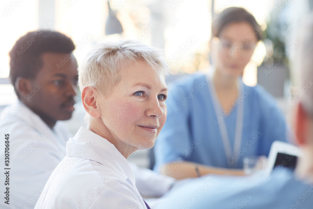 Head and shoulders portrait of mature female doctor listening to colleagues during medical council in conference room, copy space Head and shoulders portrait of mature female doctor listening to colleagues during medical council in conference room, copy space