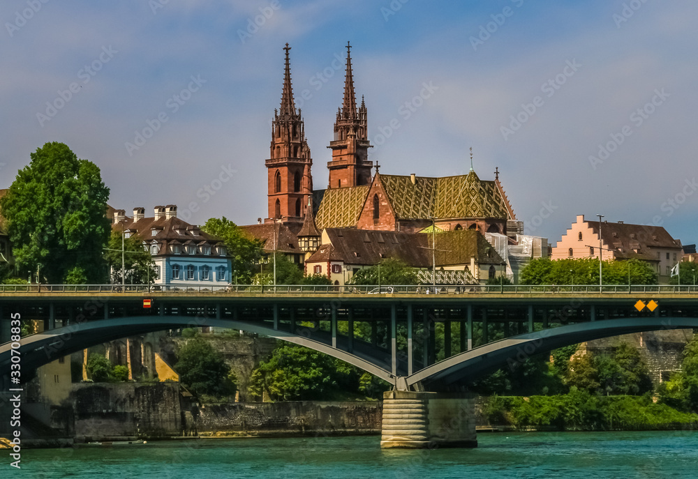 Fototapeta premium Lovely view of the Basler Münster cathedral and the bridge Wettsteinbrücke from the Rhine river. With its red sandstone walls, colourful roof tiles and twin towers, it adorns the cityscape of Basel.