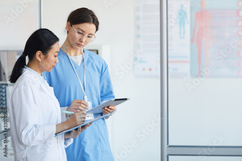 Side view portrait of two female medics filling forms while standing in medical office interior, copy space