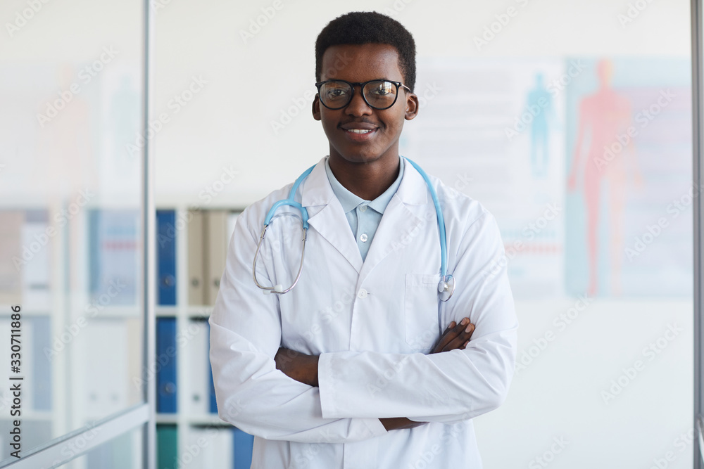 Waist up portrait of young African-American doctor standing with arms crossed and smiling at camera while posing in med clinic, copy space Waist up portrait of young African-American doctor standing with arms crossed and smiling at camera while posing in med clinic, copy space