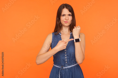 Worried brunette woman in denim dress being punctual, looking anxious and impatient about late hour, delay or deadline, pointing to wristwatch, showing clock. studio shot isolated on orange background photo