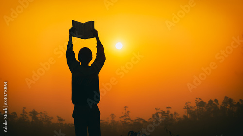 Human lift Bible up to sky with light sunset background, christian silhouette concept.