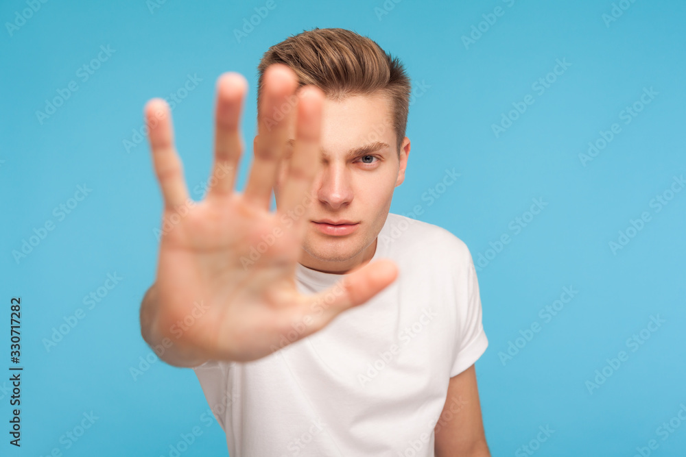 Ban, prohibition concept. Portrait of serious man in white t-shirt showing stop hand gesture, meaning caution to avoid danger or mistake, warning sign. indoor studio shot isolated on blue background Ban, prohibition concept. Portrait of serious man in white t-shirt showing stop hand gesture, meaning caution to avoid danger or mistake, warning sign. indoor studio shot isolated on blue background