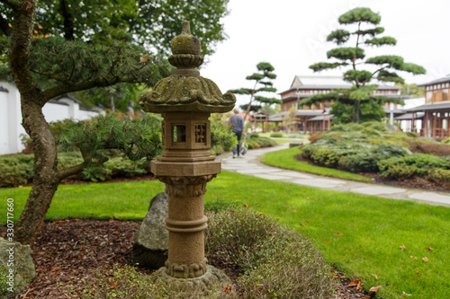 asian antique stone lantern with path on which a woman walks through park with asian topiaries and solitary shrubs