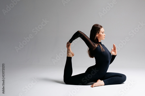 A beautiful girl of European appearance brunette sits in a difficult pose from yoga. The legs are twine, one leg is raised up, the hand holds the leg. Photo on a white background.