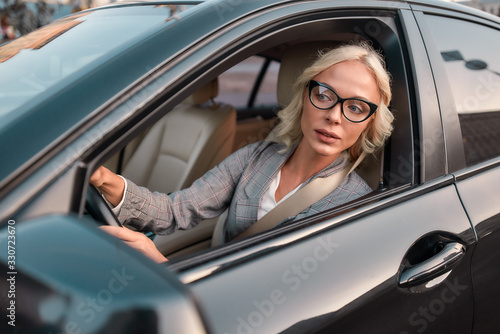 Staying in traffic jam. Portrait of young caucasian business woman in classic wear looking in the window while sitting behind the wheel of a black car © Svitlana