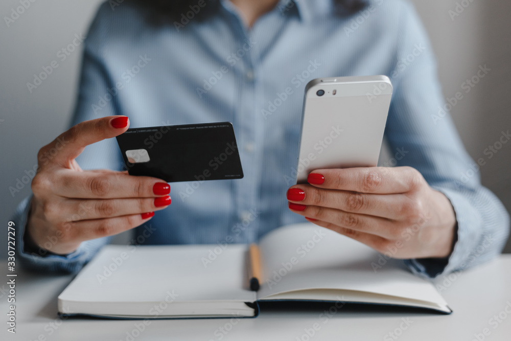 Closeup of woman's hands with red nails holding credit card and mobile cell phone making payment online wearing blue shirt. Business, freelance, self-employment. Distance job and online work concept. Closeup of woman's hands with red nails holding credit card and mobile cell phone making payment online wearing blue shirt. Business, freelance, self-employment. Distance job and online work concept.