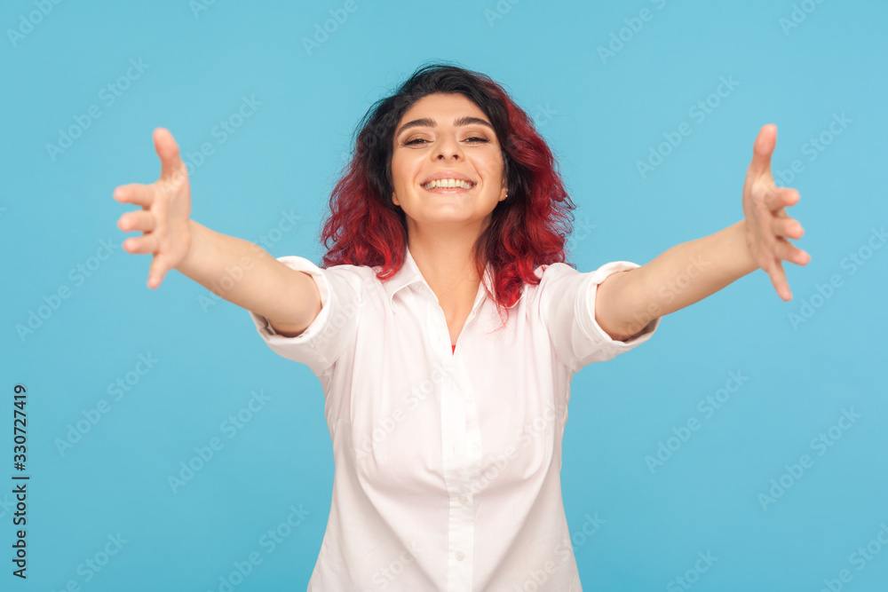 Fototapeta premium Come here, free hugs! Portrait of hipster woman with fancy red hair in shirt smiling genuinely and stretching arms to camera, welcoming ready to embrace. indoor studio shot isolated on blue background