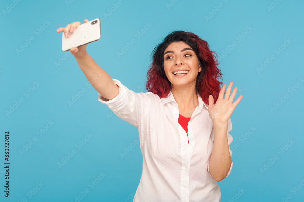 Hello, subs! Portrait of attractive hipster woman with fancy red hair waving hand saying hi while streaming on mobile phone, online blogging and communication. studio shot isolated on blue background Hello, subs! Portrait of attractive hipster woman with fancy red hair waving hand saying hi while streaming on mobile phone, online blogging and communication. studio shot isolated on blue background