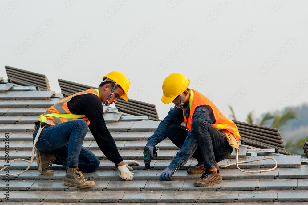 Construction worker wearing safety harness belt during working on roof structure of building on construction site,Roofer using air or pneumatic nail gun and installing concrete roof tile on top roof. - obrazy, fototapety, plakaty 