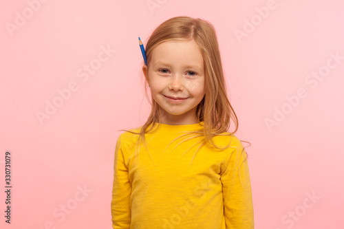 Portrait of charming adorable little girl with ginger hair holding pencil behind ear and smiling to camera, development of creative child abilities. indoor studio shot isolated on pink background