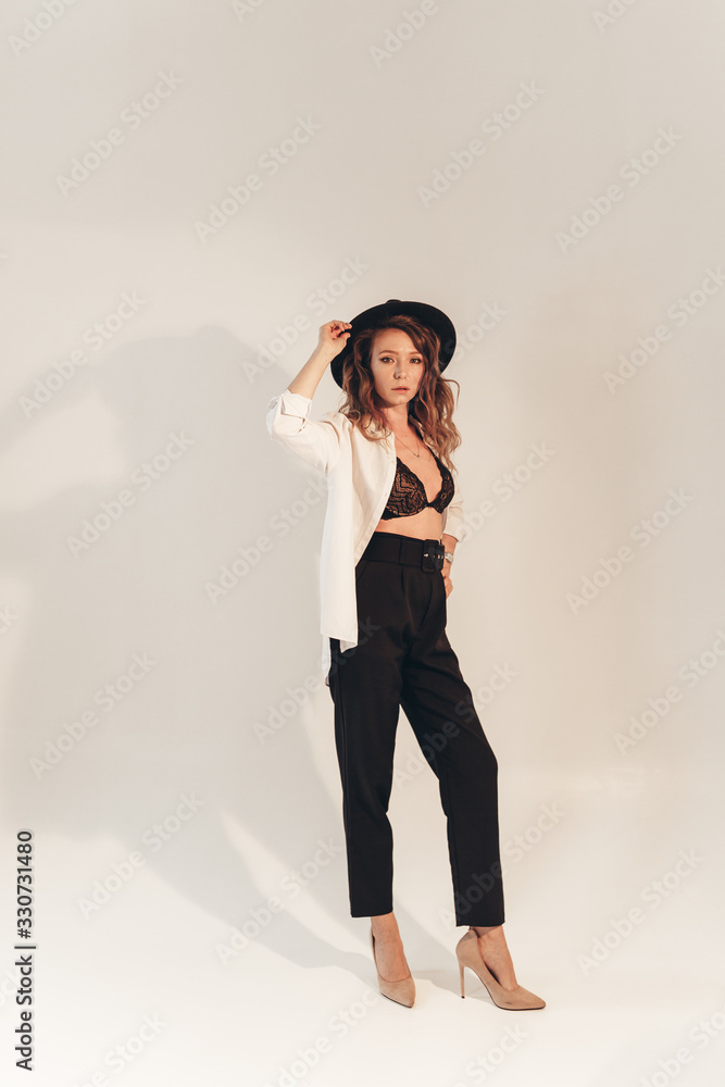 The concept of a trendy modern fashion trend shooting in a photo Studio , a beautiful sexy attractive gentle daring girl in a black hat, in black underwear, in a large shirt posing on a white backgrou The concept of a trendy modern fashion trend shooting in a photo Studio , a beautiful sexy attractive gentle daring girl in a black hat, in black underwear, in a large shirt posing on a white backgrou